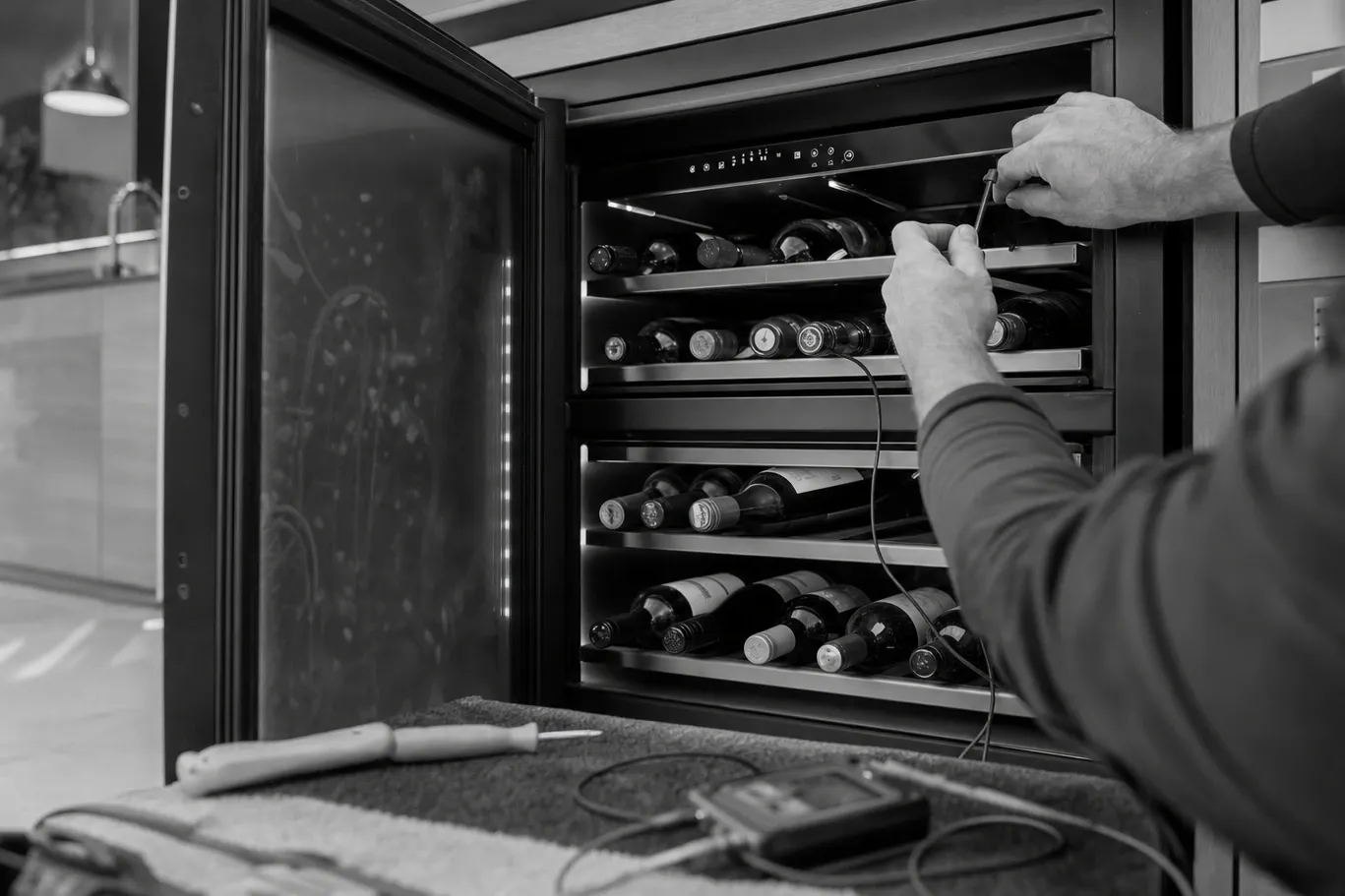 Black-and-white view of temperature probes run into an open undercounter wine cooler, bottles on the racks and a data logger on the towel below